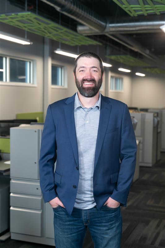 A bearded man in a blue blazer and gray shirt smiles in a modern office, representing Dynamic Lifecycle Innovations’ tech solutions.