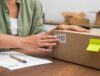 A person affixes a barcode return label to a cardboard box while preparing a product for return, with a sticky note labeled "RETURN" and paperwork on the table.