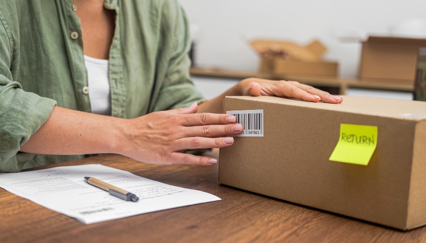 A person affixes a barcode return label to a cardboard box while preparing a product for return, with a sticky note labeled "RETURN" and paperwork on the table.