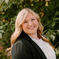 A smiling woman with long blonde hair in a dark blazer stands before green plants, representing Dynamic Lifecycle Innovations’ operations in electronics recycling services.